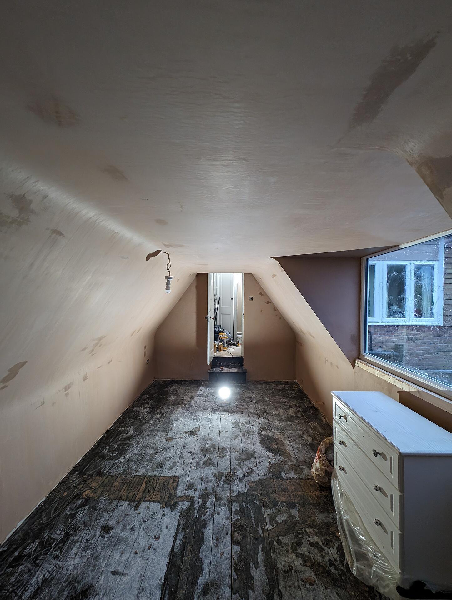 Attic bedroom with newly plastered sloping ceilings and walls, lit by a work lamp on the bare boards