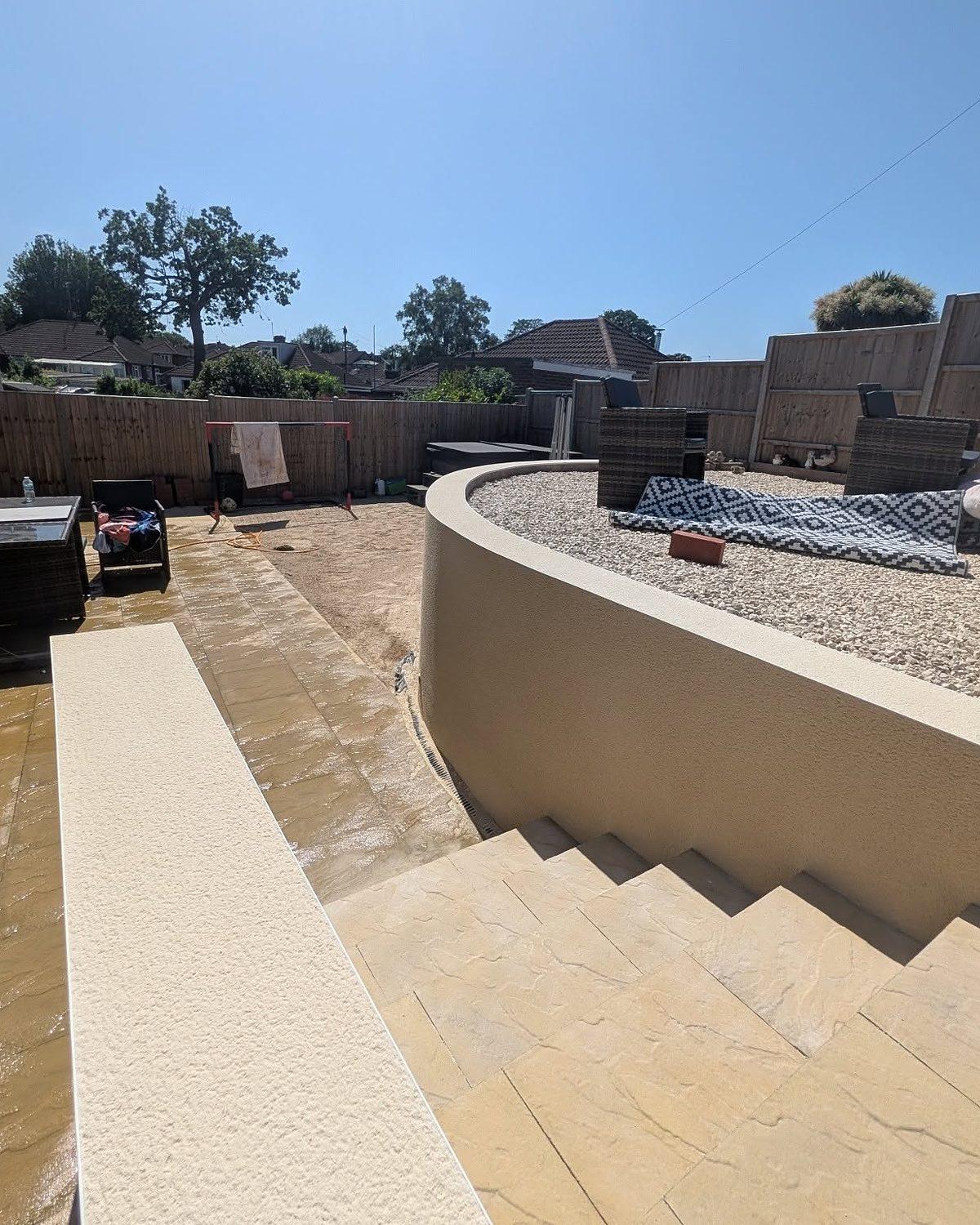 Curved garden retaining wall finished in cream render, beside new sandstone paving and steps in bright sunshine